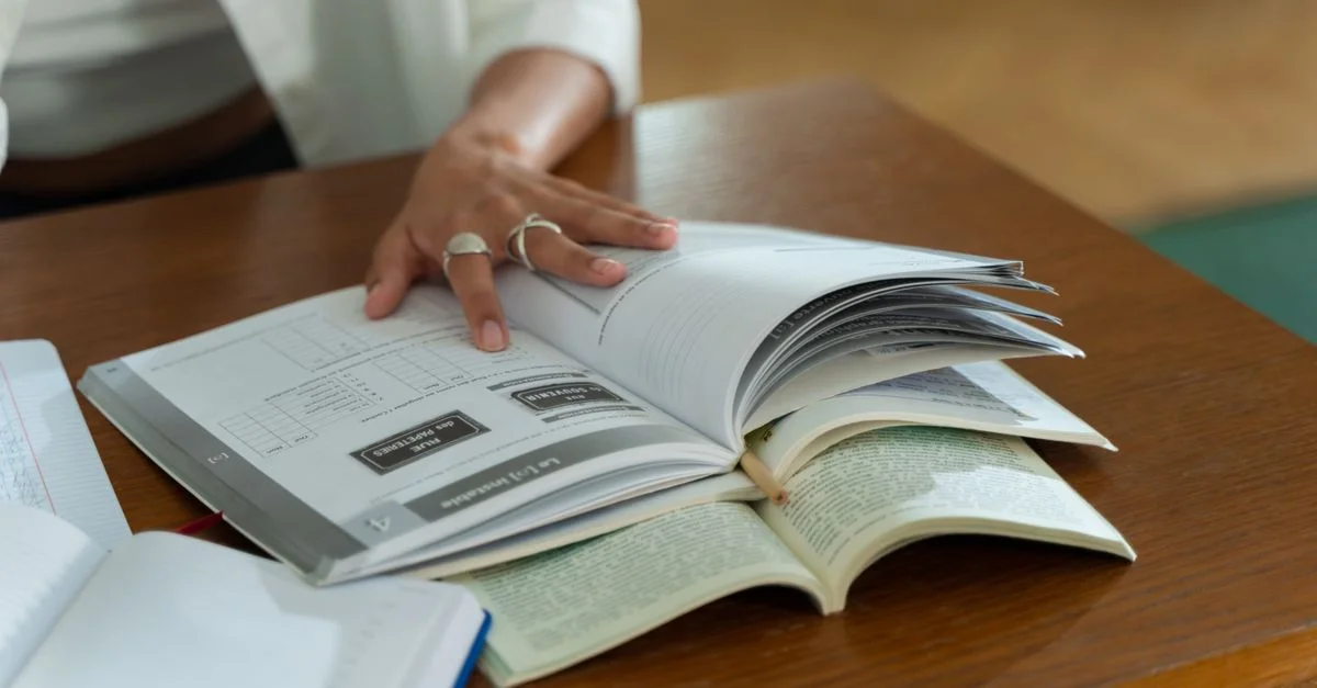 Une personne étudie avec des livres ouverts sur une table en bois, se concentrant sur le matériel écrit.